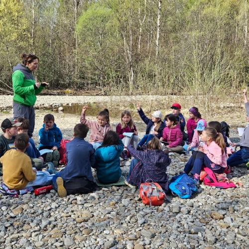 Kinder sitzen am Bach und hören der Rangerin zu