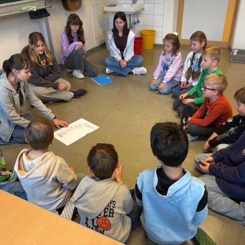 Kinder der 2a-Klasse und Schülerinnen der MS sitzen am Boden im Kreis