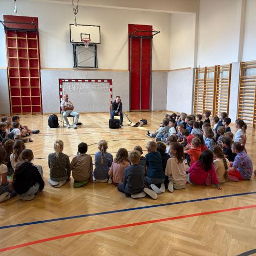 Kinder sitzen im Halbkreis auf dem Boden, vor ihnen sitzen 2 Musiklehrer