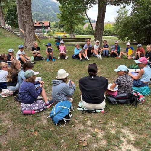 KInder und Lehrerin sitzen in einem Kreis auf der Wiese im Gras