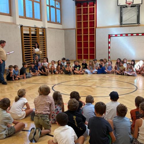 Kinder unserer Schule sitzen im Turnsaal im Kreis auf dem Boden, Schulleiter und Lehrerin stehen dazwischen