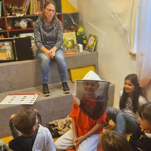 Kinder und Büchereimitarbeiterin, ein Kind trägt Imkerhaube auf dem Kopf