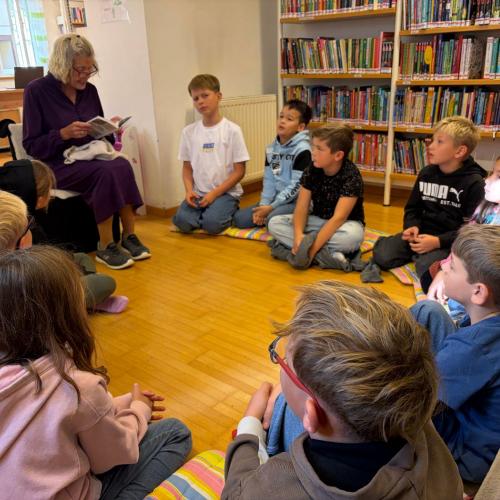 Kinder sitzen im Halbkreis, Büchereimitarbeiterin liest aus Buch vor